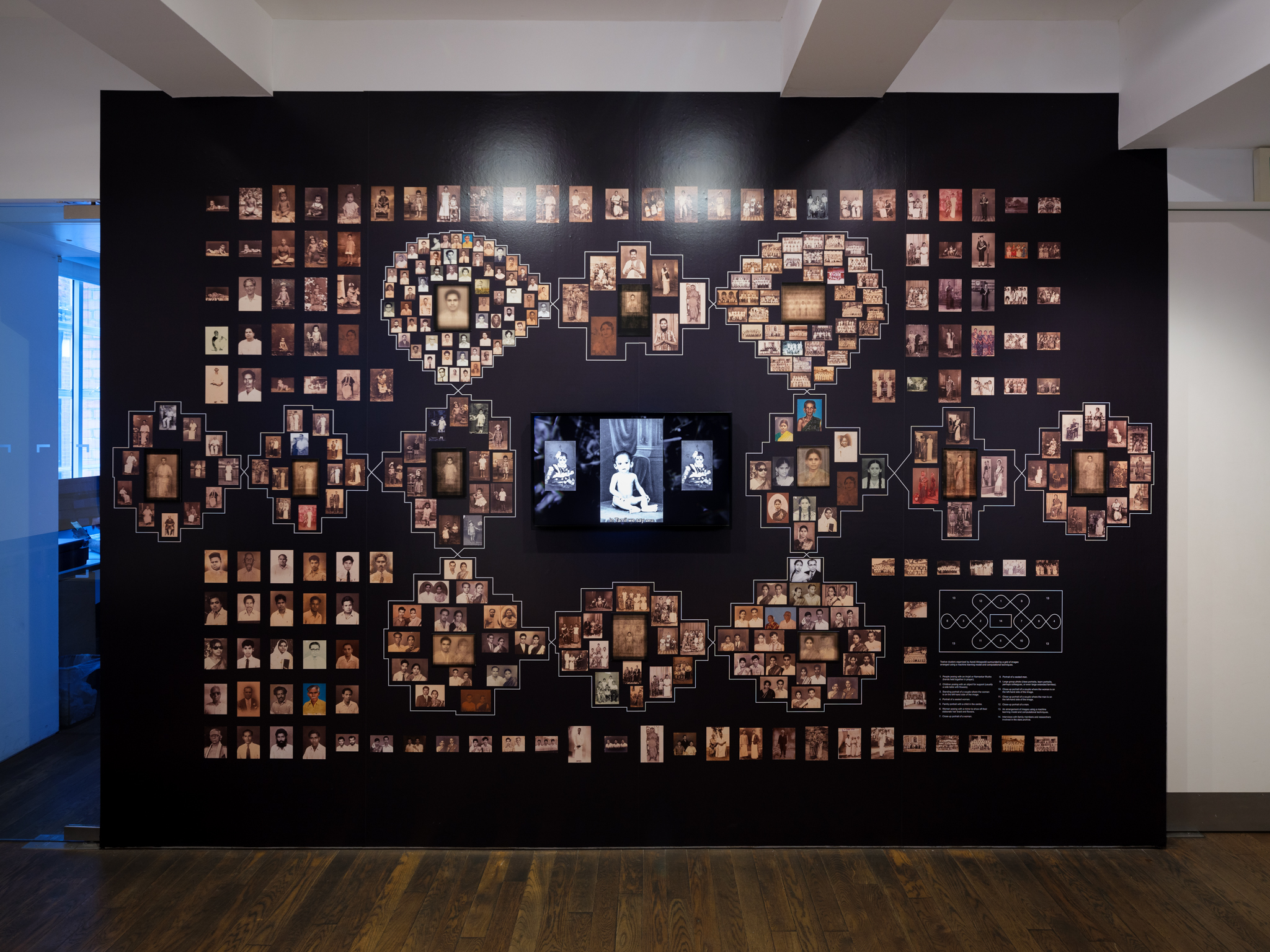 installation view: a large black wall with photographs arranged in a kolam pattern around a video screen.